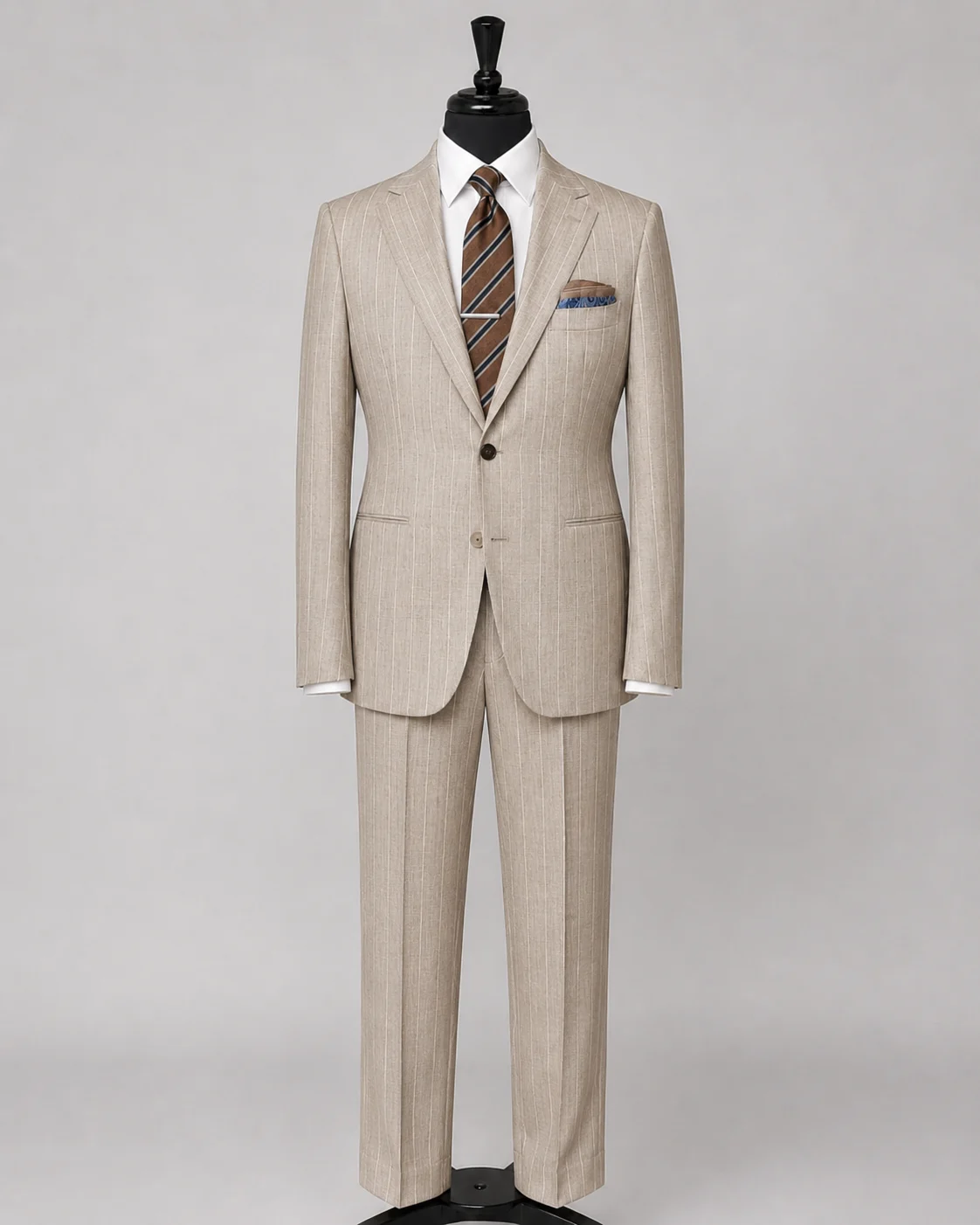 Oatmeal beige pinstripe suit displayed on a black mannequin with white shirt, brown striped tie, tie bar, and blue accent pocket square.